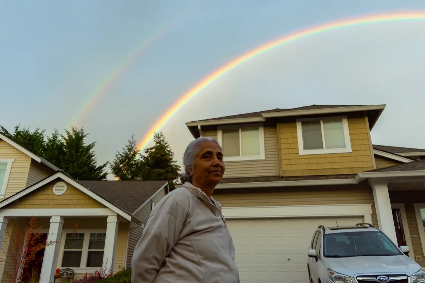 A woman standing beneath a double rainbow in a neighborhood at dusk