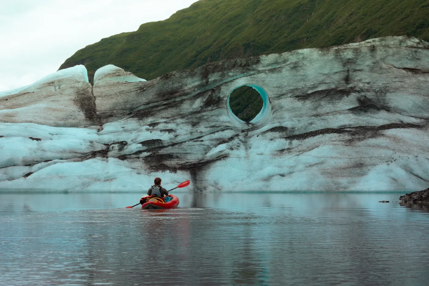 A kayaker approaching a circular opening in the face of a blue glacier