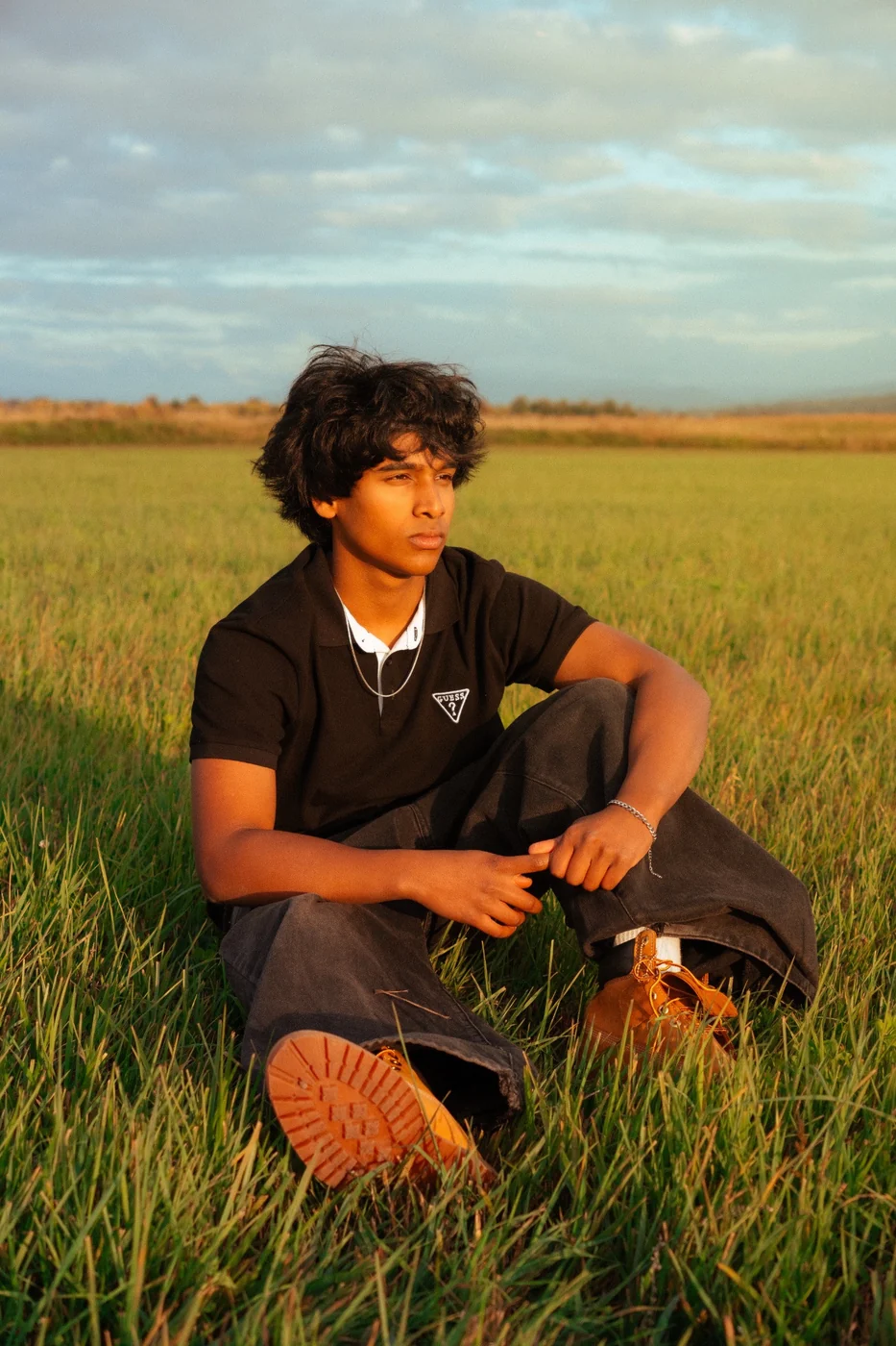 A person dressed in black sitting in tall green grass under a cloudy sky