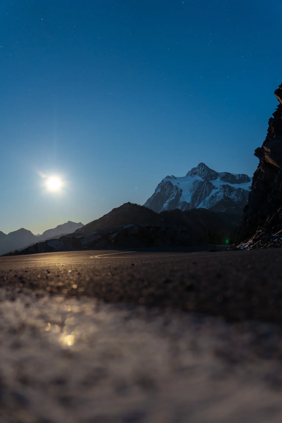 A bright moon above dark mountain ridges and a snow-covered peak under a blue night sky